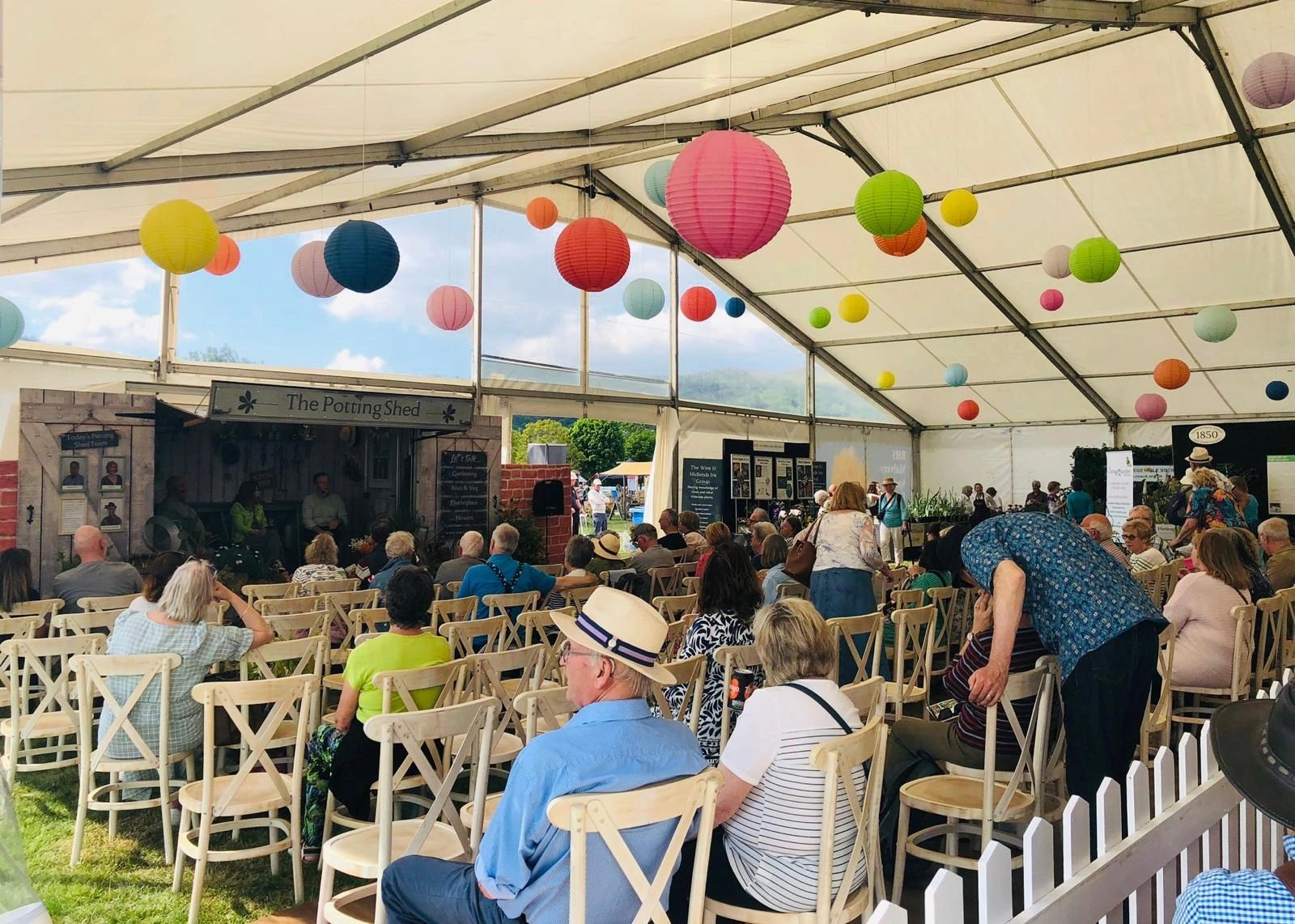 A stage titled the potted shed at a spring show, inside a marquee with an audience listening to speakers. 