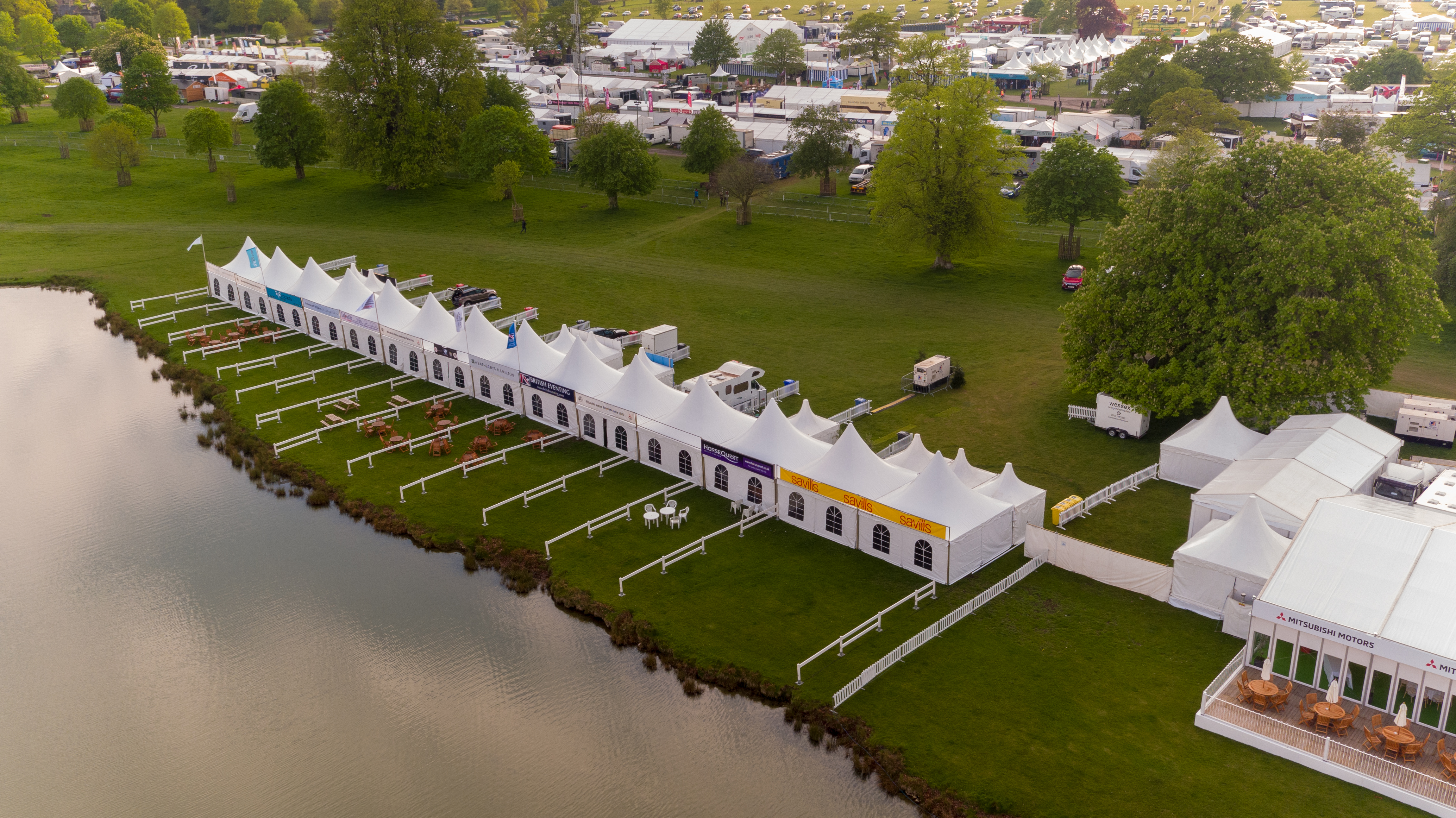 Fourteen Hat marquees are lined up in a straight line across the lake at Badminton Horse Trials. Behind the structures are lush green grass, trees and other marquees for the annual horse trials event.