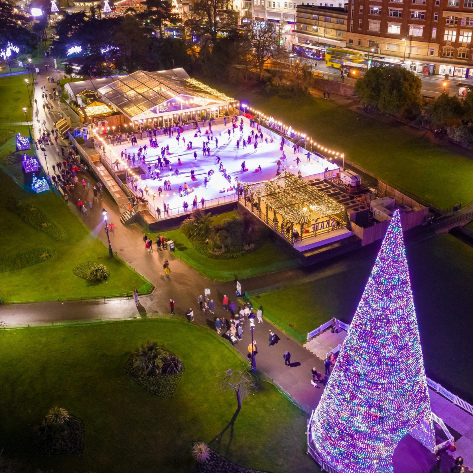 Ariel shot of Bournemouth Skate Ice Rink at night, with skaters enjoying the real ice rink, a large lit up Christmas tree and bar marquee in Bournemouth Lower Gardens. 