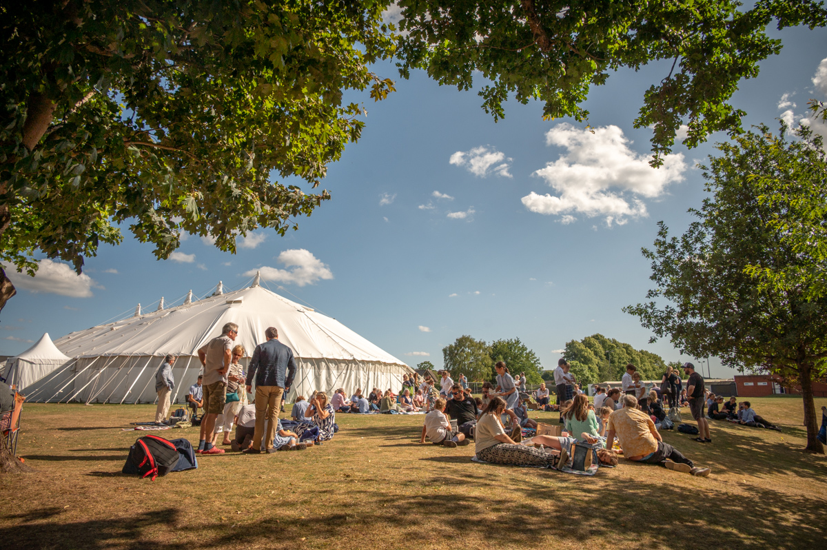 People enjoying a picnic in the summer sun, surrounded by trees and a traditional marquee at Wellington School in Somerset