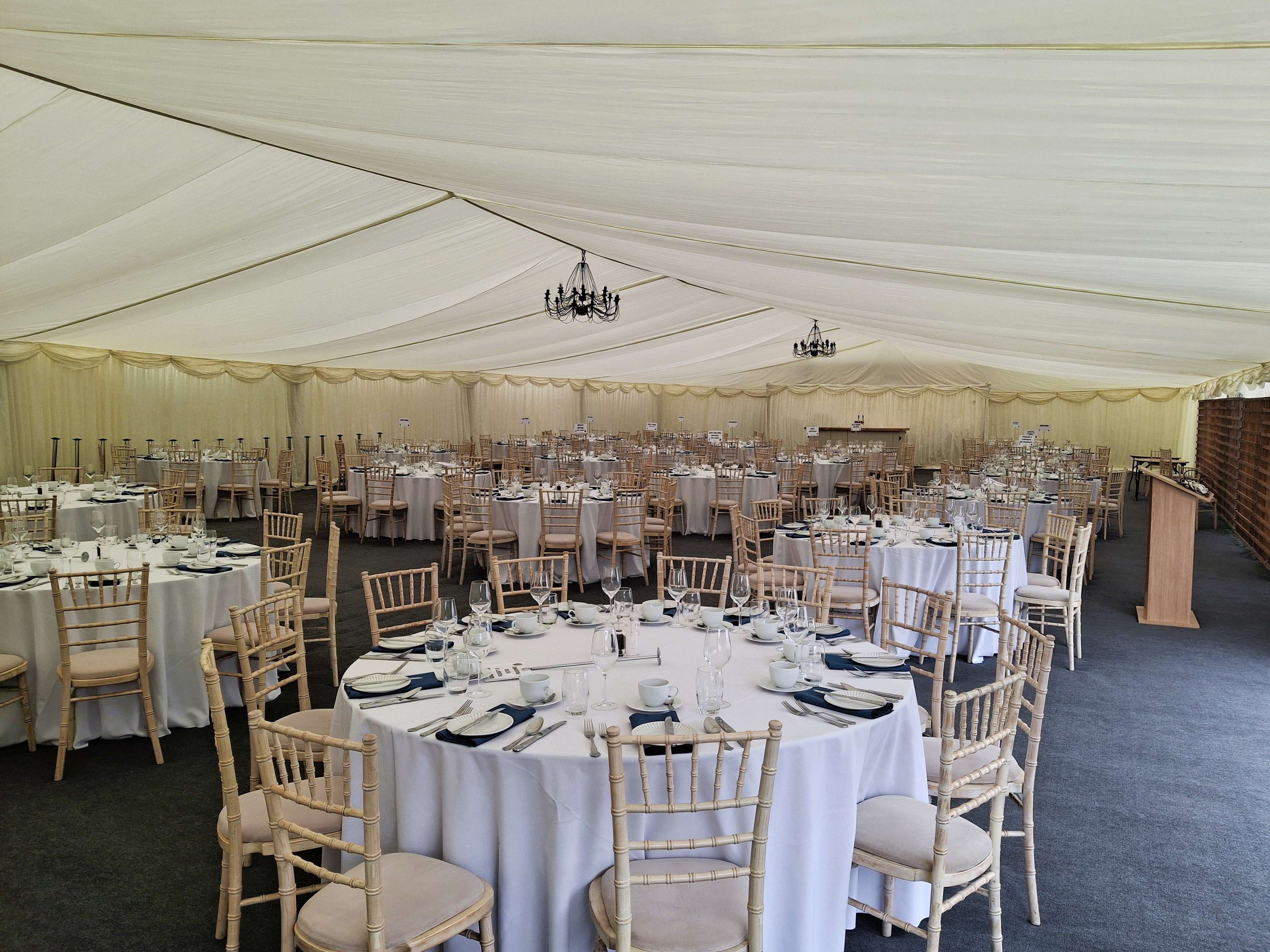 round tables with white table cloths and cutlery inside a carpeted marquee for a hospitality event. 