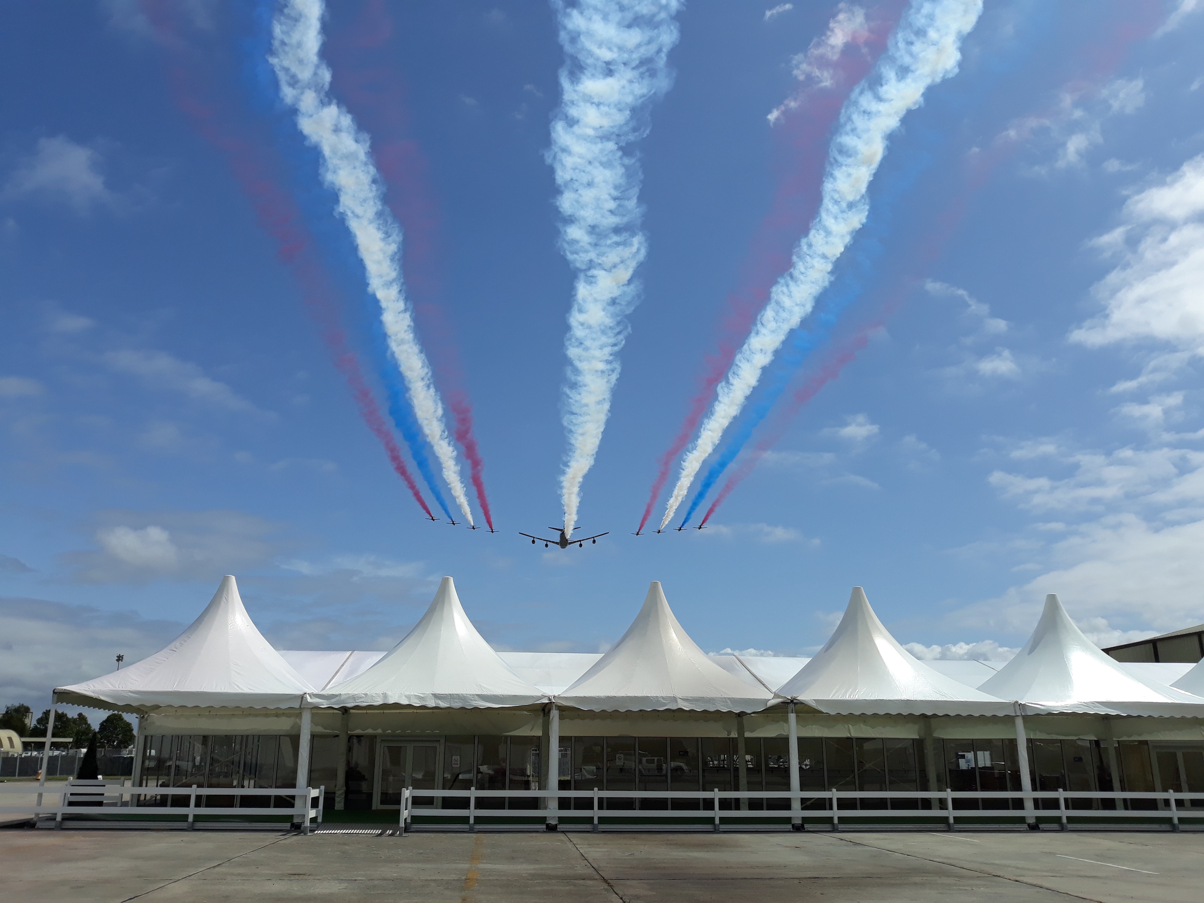 A fly past of one large plane and 4 smaller planes either side releasing red, blue and white smoke going over 5 coned shaped marquees at Fairford. 