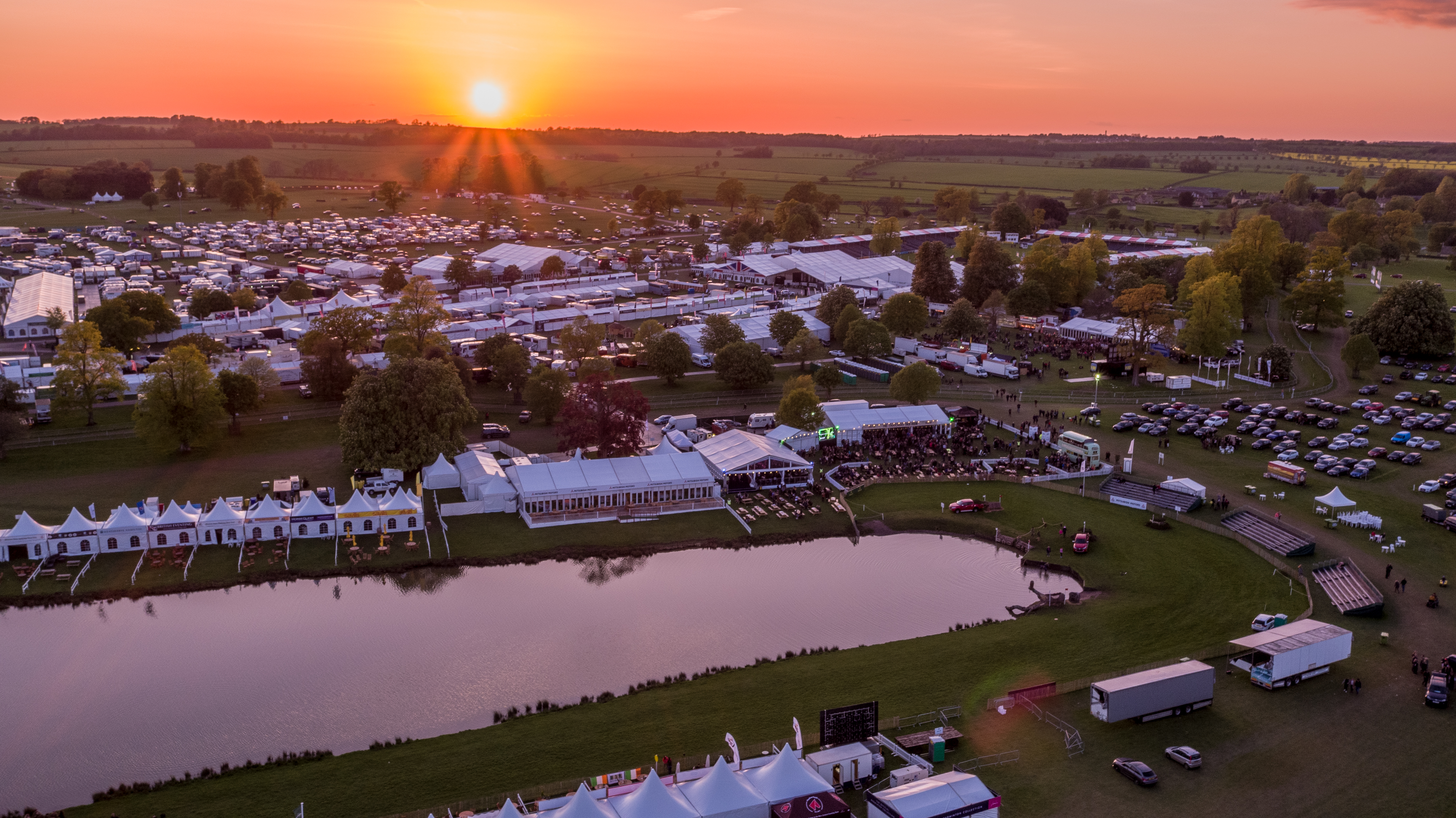 An arield view at dusck of Badminton Horse Trials. The large lake is shown at the front, with the main arena in the background. The sunsets over the fields and the event. 